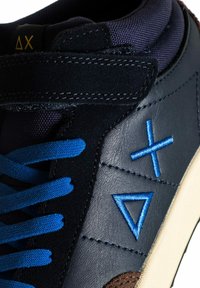 Sneaker in pelle nera con lacci blu e designs geometrici blu ricamati. Presenta una linguetta imbottita e dettagli in suede vicino alla suola.
