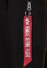 Pantaloni neri con una cerniera dotata di un cursore argentato e un'etichetta in tessuto rossa in evidenza che recita "RIMUOVERE PRIMA DEL VOLO" in lettere bianche.