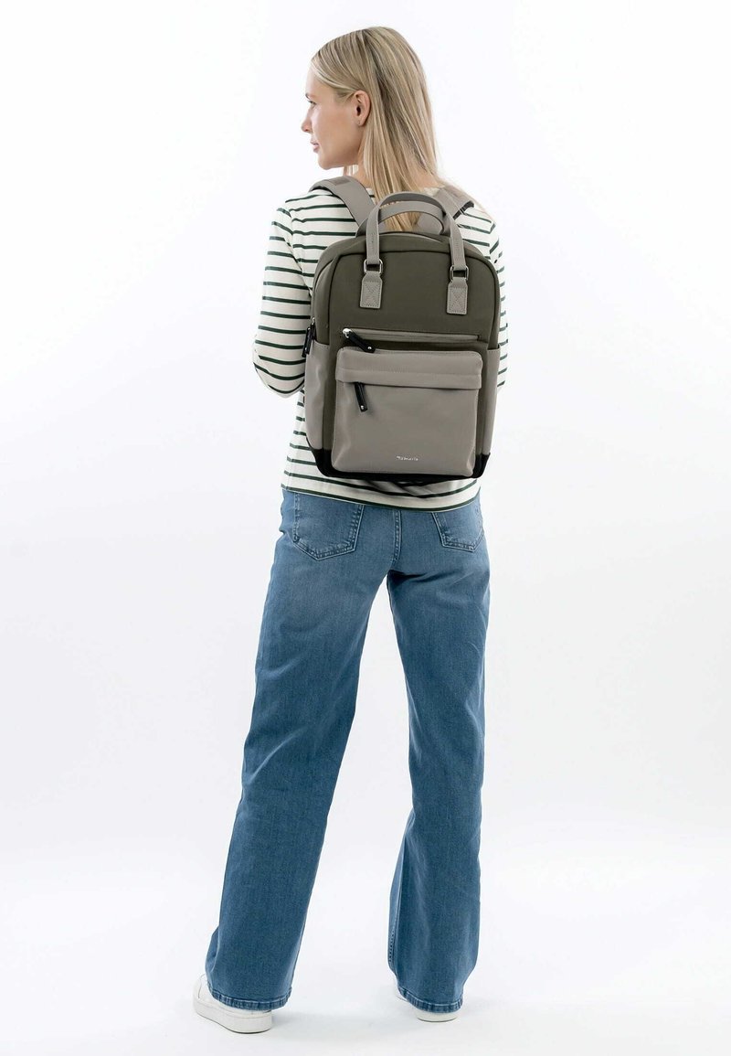 Sac à dos gris et vert avec des bretelles rembourrées, une poche zippée à l'avant, des accents en tissu et un design structuré. Haut rayé et jean bleu visibles.
