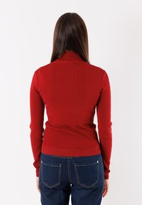 Maglione rosso a coste con colletto alto e design aderente, abbinato a jeans blu scuro con tasche posteriori e cuciture minime.