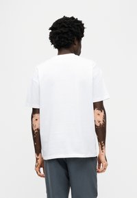 Λευκό βαμβακερό t-shirt με κοντά μανίκια, στρογγυλή λαιμόκοψη και άνετη γραμμή, εμφανιζόμενο από πίσω. Διαθέτει υφασματικά μοτίβα που μοιάζουν με υφή δέρματος στα μανίκια.