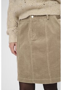 Beige Cordrock mit Gürtelschlaufen, getragen mit einem beigen Strickpullover und schwarzen Strumpfhosen, die Hand am Körper.