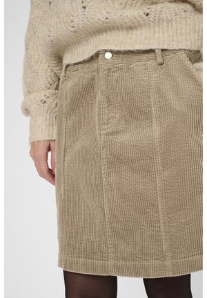 Beige Cordrock mit Gürtelschlaufen, getragen mit einem beigen Strickpullover und schwarzen Strumpfhosen, die Hand am Körper.
