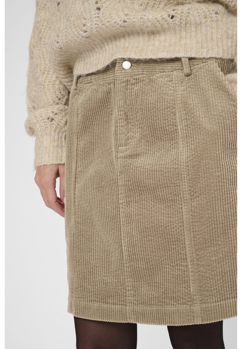 Beige Cordrock mit Gürtelschlaufen, getragen mit einem beigen Strickpullover und schwarzen Strumpfhosen, die Hand am Körper.