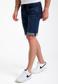 Shorts de mezclilla en tono oscuro con puños enrollados; presenta un clásico diseño de cinco bolsillos y costuras sutiles. Combinados con zapatillas Nike blancas.