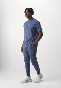 Niebieska sportowa koszulka z czarnym logo na piersi, zestawiona z pasującymi niebieskimi dresami i białymi trampkami. Gładki materiał, luźny krój.