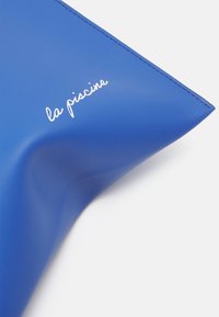 Niebieska, strukturalna torba wykonana z gładkiego materiału, z białym napisem "la piscine". Projekt ma kanciaste kształty i eleganckie wykończenie.