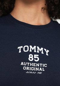 Námornícka modrá rebrovaná tričko s okrúhlym výstrihom. Biele potlačené texty hovoria „TOMMY 85 AUTHENTIC ORIGINAL since 1985.“ Mäkká štruktúra, neformálny štýl.