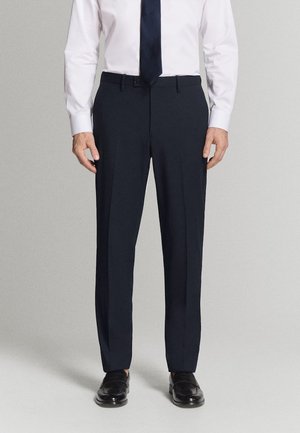 Mężczyzna ubrany w granatowe spodnie garniturowe, białą koszulę, czarny krawat oraz czarne skórzane buty, stojący na prostym jasnoszarym tle.