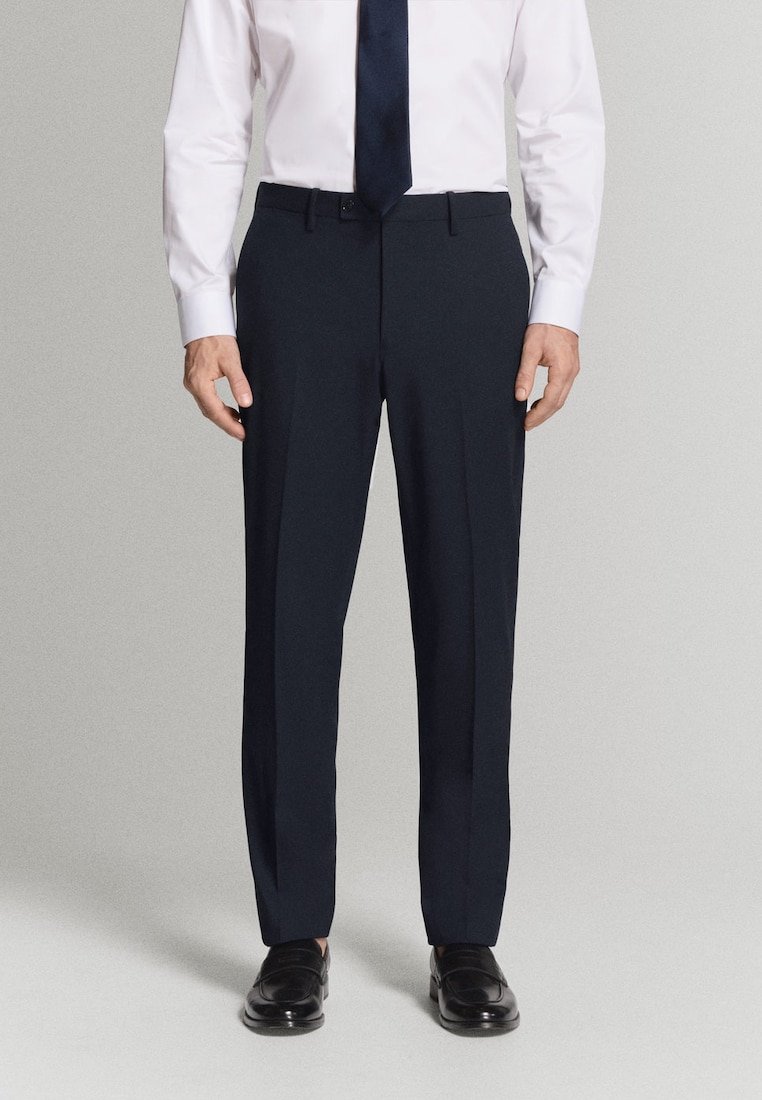 Mężczyzna ubrany w granatowe spodnie garniturowe, białą koszulę, czarny krawat oraz czarne skórzane buty, stojący na prostym jasnoszarym tle.