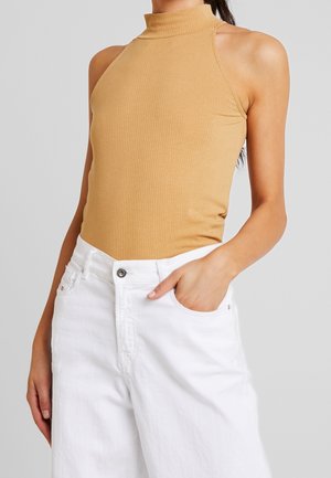 Femme portant un haut col roulé sans manches côtelé de couleur camel, rentré dans un jean en denim blanc taille haute, main dans la poche.