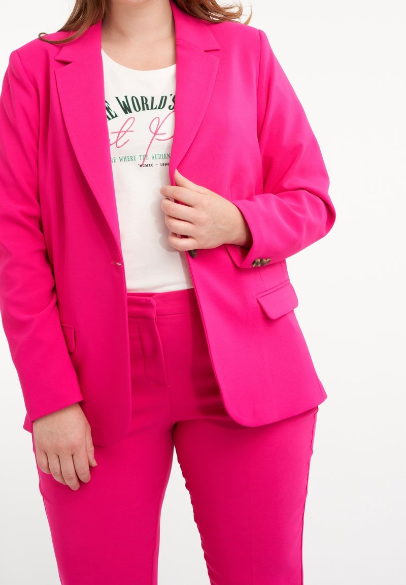 Blazer rosa brillante con pantalones a juego, combinado con una camiseta blanca con texto verde. El blazer tiene solapa entallada y un cierre de un solo botón.
