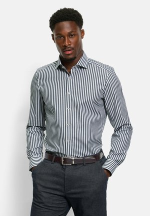 Blauw-wit gestreepte overhemddress met een button-down kraag en een getailleerd silhouet, gecombineerd met donkere broeken en een leren riem in het bruin.