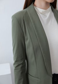 Olivově zelený blejzr s přiléhavým střihem, přední záhyby, decentní ozdobné prvky a hladkou texturu, v kombinaci s bílým topem s vysokým límcem.