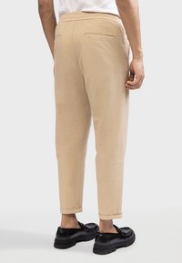 Pantalons beiges à coupe décontractée, avec une taille élastique et deux poches arrière. Le tissu semble lisse avec une texture subtile, retroussé aux chevilles.