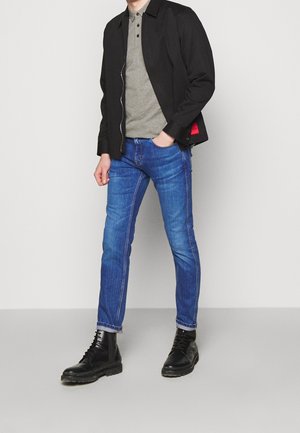 Chaqueta negra, camiseta polo gris, jeans azules con etiqueta roja y botas de cordones negras. El conjunto presenta un diseño moderno y casual.