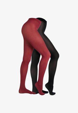 Frau trägt schwarzen BH, transparente schwarze Strumpfhosen und schwarze High Heels, steht mit überkreuzten Beinen vor einem schlichten weißen Hintergrund.