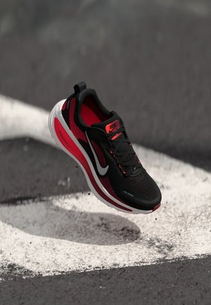 Black and red running shoe featuring a mesh upper, textured pattern, cushioned sole, and a white swoosh logo. Floating above a painted track line.