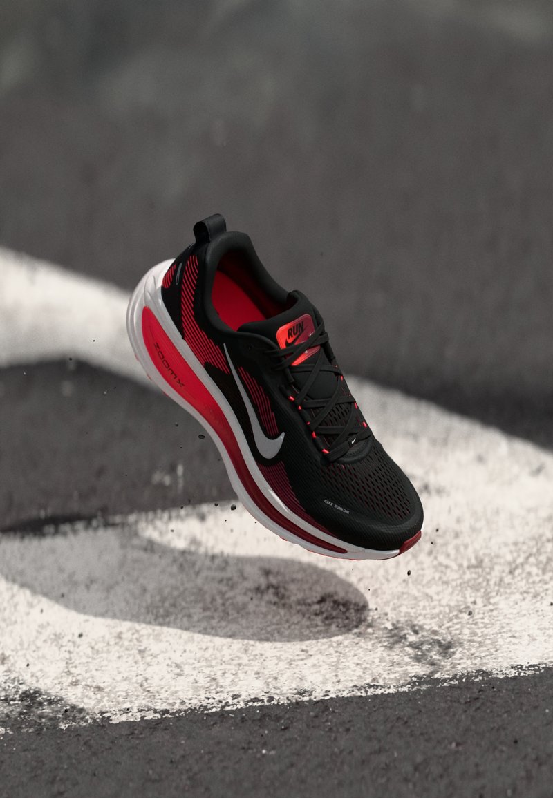 Chaussure de course noire et rouge avec un dessus en maille, un motif textur�é, une semelle rembourrée et un logo swoosh blanc. Flottant au-dessus d'une ligne de piste peinte.