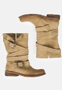 Botas de tobillo de ante beige con múltiples correas cruzadas, un detalle de hebilla y un tacón de madera bajo. Superficie texturizada con punta redondeada.