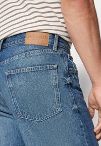 Jeans din denim albastru cu un finisaj texturat, având o etichetă din piele maro și buzunare la spate, prezentate din lateral.