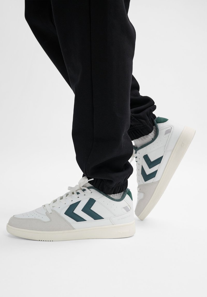 Person trägt weiße Sneakers mit dunkelgrünen Chevron-Streifen und schwarze Hose vor einfachem hellem Hintergrund.