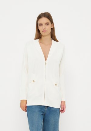 Donna che indossa un cardigan bianco con zip e dettagli di bottoni dorati sopra jeans blu, in piedi davanti a uno sfondo bianco uniforme.