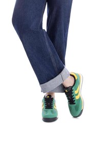 Zapatillas deportivas verdes y amarillas con cordones negros, parte superior texturizada y suela blanca. Llevadas con pantalones de mezclilla oscuros con dobladillo enrollado.