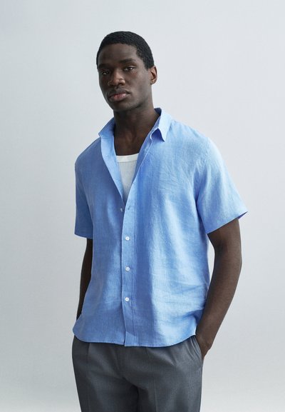 Jeune homme portant une chemise à manches courtes bleu clair boutonnée par-dessus un débardeur blanc, avec un pantalon gris, debout les mains dans les poches.