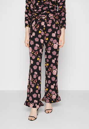 Czarne spodnie o szerokich nogawkach z różowymi, niebieskimi i żółtymi kwiatowymi wzorami oraz falbankami na dole, noszone z czarnymi sandałami na wysokim obcasie z paskami.