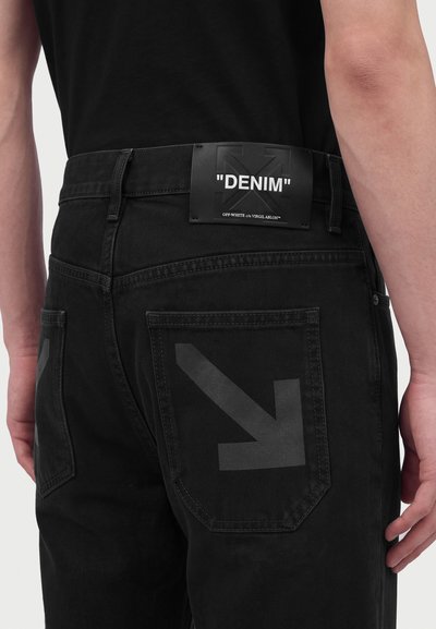 Μαύρο τζιν με δερμάτινη ετικέτα που γράφει "DENIM" και μεγάλες σκοτεινές βέλη τυπωμένες στις πίσω τσέπες.