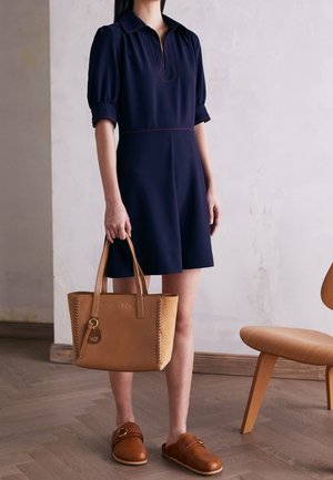 Marineblauwe jurk met korte mouwen en een kraag, met contrasterende rode stiksels; gecombineerd met een beige leren handtas en bruine instapschoenen.