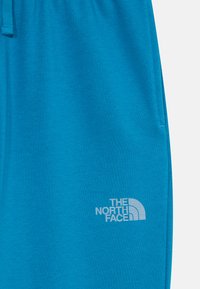 Mėlyni sportiniai džinsai, pagaminti iš minkštos medžiagos, su elastingu juosmeniu ir šonine kišene, ant šlaunies esantis baltas "The North Face" logotipas.