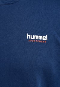 Marineblauer Stoff mit weißem „hummel“-Logo über kleinerem orangem Schriftzug „SPORTSWEAR“ auf einem strukturierten Sportbekleidungsstück.