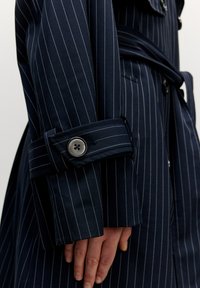 Marinblå trenchcoat med rutigt mönster och en strukturerad design. Har en manschett med en knapp och ett knytbälte, vilket visar på en slät tygtextur.