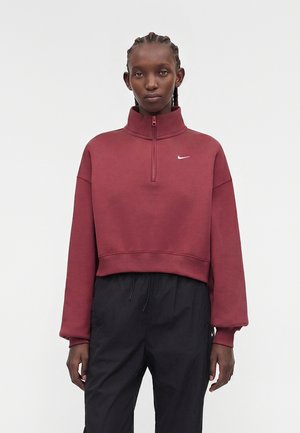 Femme aux cheveux tressés portant un sweat Nike bordeaux à demi-fermeture éclair et un pantalon noir à cordon, debout devant un fond uni.