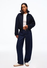 Nainen, joka pukeutuu tummansiniseen lyhytpukuiseen takkiin, valkoiseen vartalonmyötäiseen lyhytpaitaan, leveälahkeisiin tummiin farkkuihin ja beigen värisiin tennareihin, seisoo valkoista taustaa vasten.