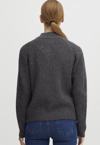 Persona con coda di cavallo castano chiaro, che indossa un maglione a coste grigio scuro e jeans blu, in piedi di spalle contro uno sfondo chiaro e semplice.