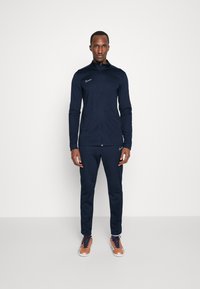 Marineblauw sporttracksuit gemaakt van rekbare stof, met een ritsjack en tapered broek, vergezeld van Nike-logo details en lichte schoenen.