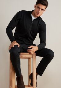 Svart, zippered sweatshirt med långa ärmar, tillverkad av mjukt tyg. Bärs med matchande svarta byxor och bruna ankelkängor på en träpall.