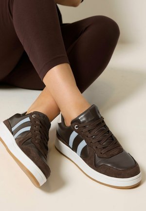 Persoon met donkerbruine sneakers met lichtblauwe strepen en witte zolen, benen gekruist, gekleed in donkerbruine leggings, zittend op de grond.