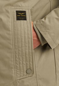 Beige jacka med en ribbad ficka och en gyllene logotypplåt. Slät tyg, synliga sömmar och en rund metallaccent fullbordar designen.