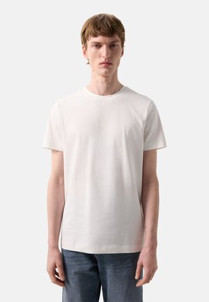 Młody mężczyzna w prostej białej koszulce z krótkim rękawem z małym wypukłym logo na piersi, w połączeniu z niebieskimi dżinsami, stojący na białym tle.