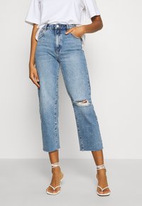 Ljusblå högmidjade jeans med fransad nederkant och stort hål vid knäet, med fem fickor och knappstängning. Bärs med vita sandaler med remmar.