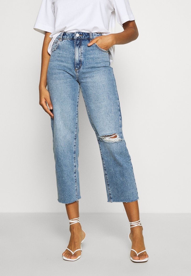 Ljusblå högmidjade jeans med fransad nederkant och stort hål vid knäet, med fem fickor och knappstängning. Bärs med vita sandaler med remmar.