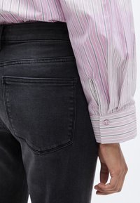 Μπλούζα με μακριά μανίκια σε ανοιχτό ροζ χρώμα με κατακόρυφες λευκές ρίγες, συνδυασμένη με στενά εφαρμοστά τζιν σε σκούρο γκρι. Διαθέτει μανσέτες με κουμπιά και δύο τσέπες στην πλάτη.