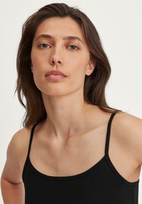 Débardeur noir à fines bretelles, présentant une coupe ajustée et une texture douce et lisse. Gros plan montrant le décolleté et les détails des épaules.