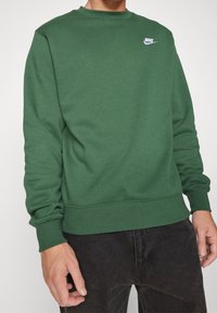 Grön sweatshirt gjord av mjukt tyg, med rundad halsringning, som har en liten vit Nike-logga på vänster bröst och ribbade mudd och nederkant.