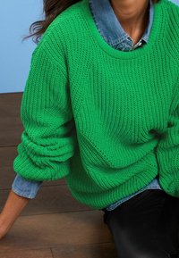 Vrouw draagt een felgroene gebreide trui over een denim shirt, zittend op een houten vloer tegen een lichtblauwe achtergrond.