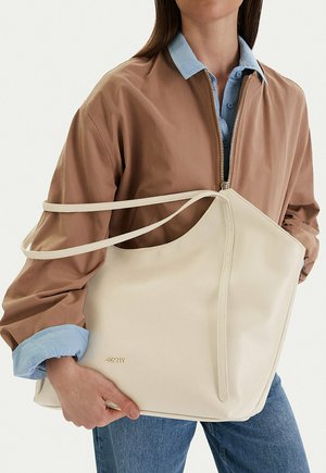 Persona con una chaqueta marrón y camisa azul sosteniendo un bolso grande beige con el logo dorado "JENNY" en la parte frontal.